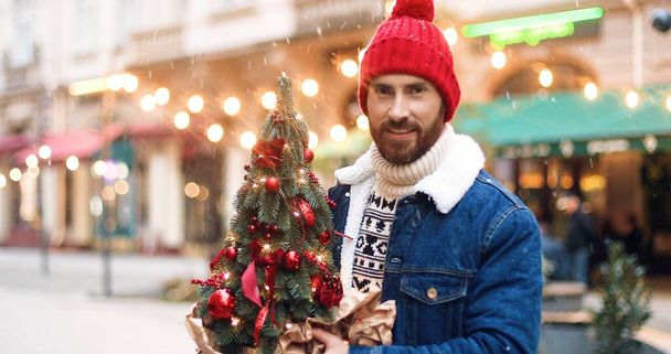 Close up of handsome happy Caucasian man in red warm hat standing in city decorated with lights with small christmas tree in hands. Portrait of guy on street on new years eve. Xmas concept - Foto, imagen