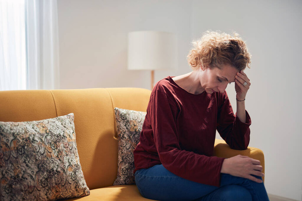 Женщина с болью в колене и головной болью сидит дома на диване. - Фото, изображение