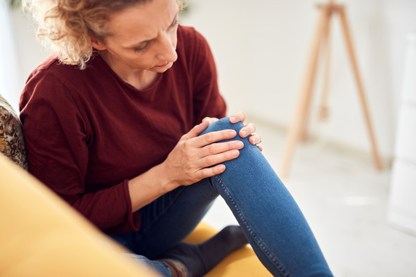 Γυναίκα με πόνο στο γόνατο / πρόβλημα κάθεται σε έναν καναπέ στο σπίτι. - Φωτογραφία, εικόνα