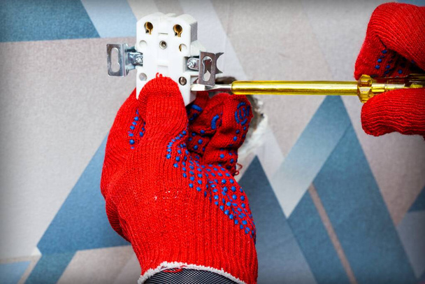 Un electricista con guantes protectores rojos utiliza un destornillador para instalar un enchufe en la pared de la sala de estar. Primer plano de las manos. Concepto maestro del hogar. - Foto, Imagen