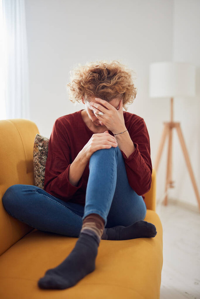 Женщина с болью в колене и головной болью сидит дома на диване. - Фото, изображение