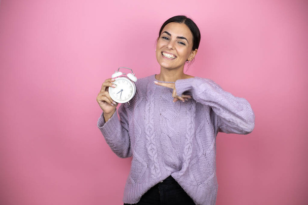 Beautiful woman wearing a casual violet sweater over pink background pointing, scared and holding a clock - Фото, зображення