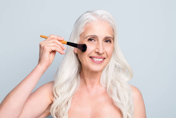 Close up photo of beautiful retired woman applying brush cheekbone powder makeup isolated grey color background - 写真・画像