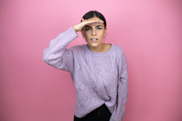 Beautiful woman wearing a casual violet sweater over pink background very happy and smiling looking far away with hand over head. searching concept - Foto, imagen