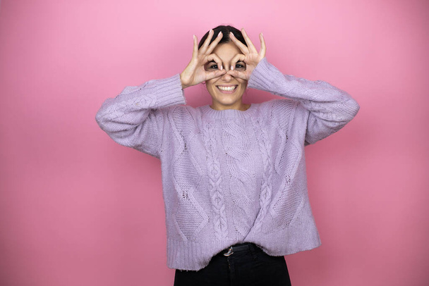 Hermosa mujer con un suéter violeta casual sobre fondo rosa haciendo buen gesto conmocionado con la cara sonriente, ojos mirando a través de los dedos - Foto, imagen