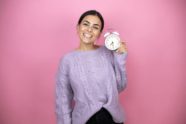 Beautiful woman wearing a casual violet sweater over pink background smiling confident holding a clock - Фото, зображення