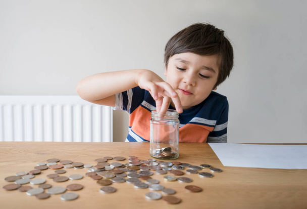 学校の子供はお金のコインを明確な瓶に入れ、子供は彼の節約のお金を数え、若い男の子は彼の手にコインを持って、子供は将来の概念のための節約について学ぶ - 写真・画像