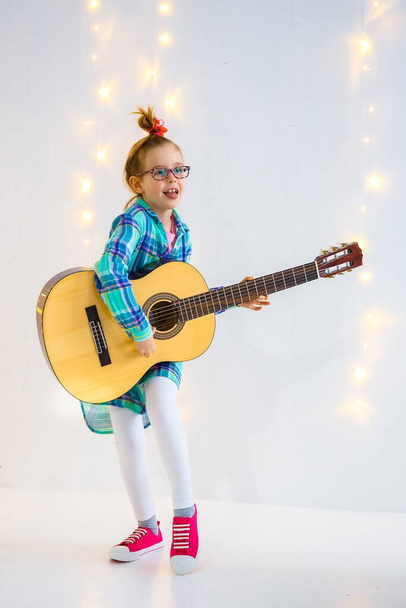 Emotional little girl with guitar in studio. Charming beauty plays the guitar with a slight smile on her face - Foto, afbeelding