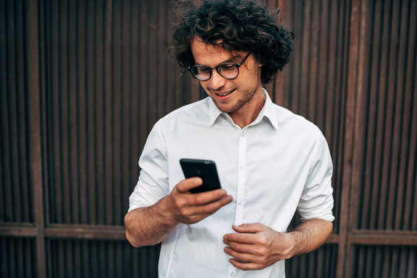 Closeup of happy businessman in white shirt with transparent eyeglasses messaging on mobile phone. Handsome curly male has online conference with a colleague via smartphone outside. - Foto, Imagen