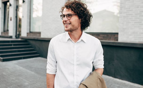 Attractive businessman in eyewear smiling broadly posing outdoors. Male entrepreneur resting in the city street. The curly guy in casual wears spectacles with curly hair walking outside after work - Foto, Imagen