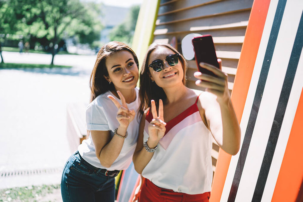 Caucasian hipster girl making together selfie photos for sharing to own web blog using smartphone front camera on leisure, happy female friends showing V-sign while shooting influence video - Фото, зображення