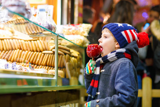 Счастливый ребенок ест на яблоке, покрытом красным сахаром на рождественской ярмарке - Фото, изображение
