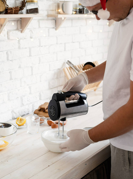 Joulun leivontakonsepti. Mies joulupukki hattu ruoskinta munanvalkuaiset kanssa sähköinen mikseri keittiössä - Valokuva, kuva