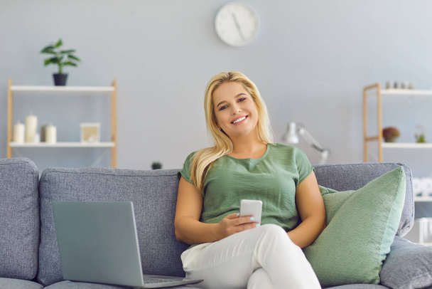 Šťastná žena sedí na gauči s laptopem, drží mobilní telefon v ruce a dívá se do kamery - Fotografie, Obrázek