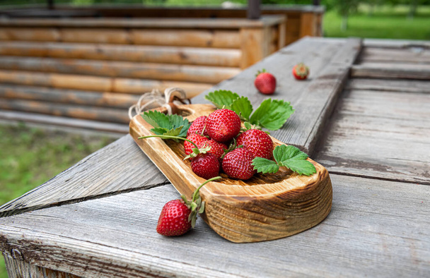 Few bright red organic strawberries on handmade wooden board and on old textured wooden surface. Unfocused real garden environment at background. Selective focus. Healthy eating and dieting concepts. - Foto, afbeelding