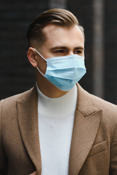 Businessman and coronavirus epidemic. Close up portrait of young business man in a disposable facial mask. A man defends himself against covid 19 on the big city street. Modern buildings at background - Fotografie, Obrázek