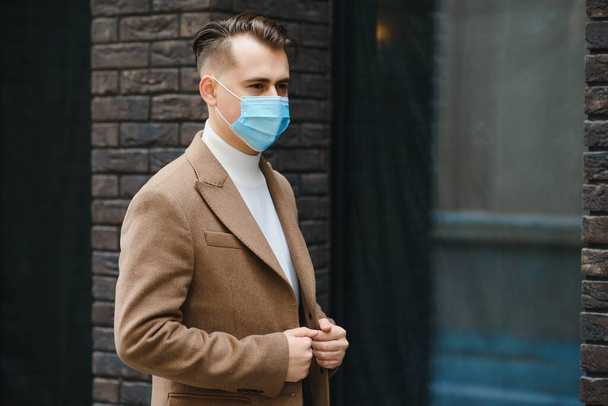 Businessman and coronavirus epidemic. Close up portrait of young business man in a disposable facial mask. A man defends himself against covid 19 on the big city street. Modern buildings at background - Foto, imagen