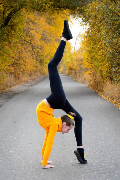 Stretching and gymnastics. A young girl in a yellow pullover stands in a gymnastic pose against a background of yellow foliage. Vertical - Fotografie, Obrázek
