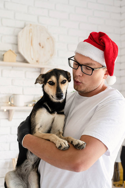Noel yemeği. Noel Baba şapkalı genç adam mutfakta köpeğini kollarında tutuyor. - Fotoğraf, Görsel