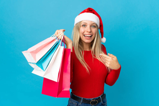 Γυναίκα με χριστουγεννιάτικο καπέλο πρόκειται να αγοράσει κάτι απομονωμένο σε μπλε φόντο με έκπληξη έκφραση προσώπου - Φωτογραφία, εικόνα