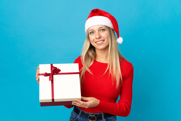 Γυναίκα με χριστουγεννιάτικο καπέλο κρατώντας ένα δώρο απομονωμένο σε μπλε φόντο χαμογελώντας πολύ - Φωτογραφία, εικόνα