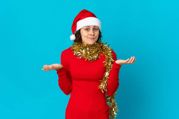 English girl with christmas hat isolated on blue background having doubts while raising hands - Photo, image