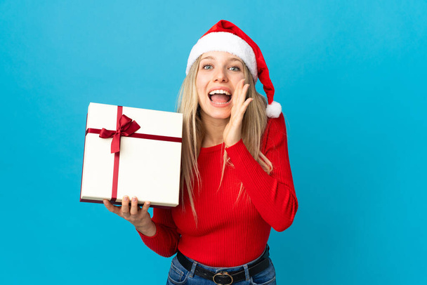 Woman with christmas hat holding a present isolated on blue background shouting with mouth wide open - Foto, immagini
