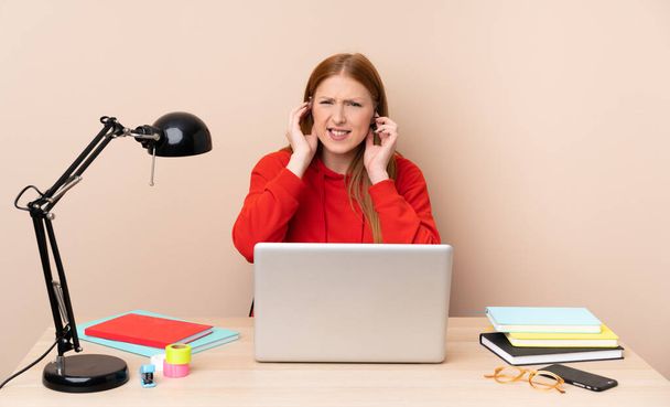 Mujer joven estudiante en un lugar de trabajo con un ordenador portátil frustrado y cubriendo oídos - Foto, imagen