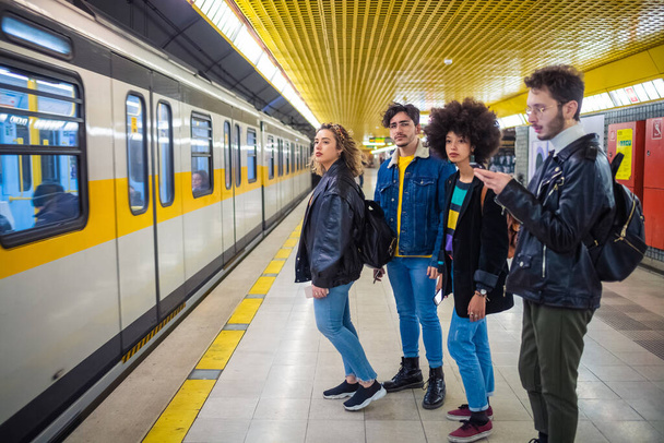 Ομάδα τεσσάρων πολυεθνικών ανθρώπων τουριστικός μετακινούμενος στο υπόγειο περιμένοντας για το μετρό χρησιμοποιώντας smartphone  - Φωτογραφία, εικόνα