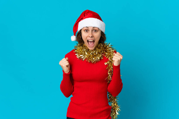 Anglická dívka s vánočním kloboukem izolované na modrém pozadí slaví vítězství v pozici vítěze - Fotografie, Obrázek