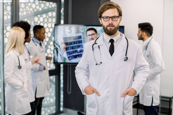 Handsome young bearded Caucasian male doctor posing to camera at modern clinic. Team of multi-ethnic doctors on the background, discussing CT results and having video call with male doctor - Фото, изображение