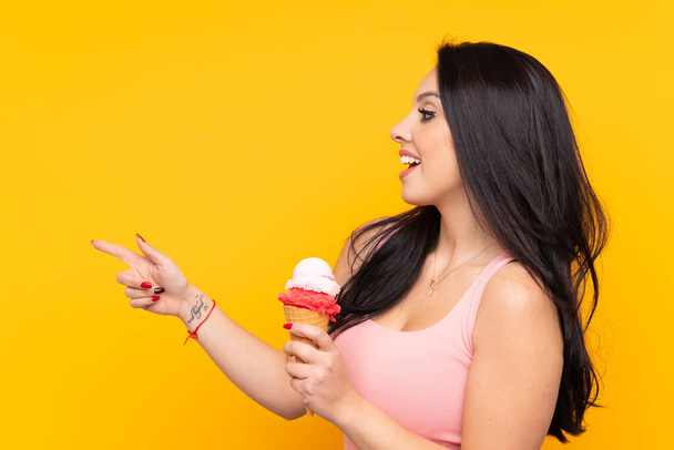 Молода колумбійська дівчина, яка тримає морозиво з корнетом над ізольованим жовтим фоном, вказує на бік, щоб представити продукт - Фото, зображення