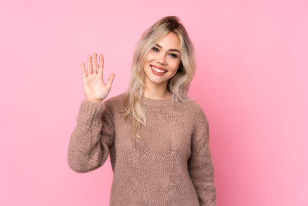 Teenager ξανθιά κοπέλα φορώντας ένα πουλόβερ πάνω από απομονωμένο ροζ φόντο χαιρετισμό με το χέρι με χαρούμενη έκφραση - Φωτογραφία, εικόνα