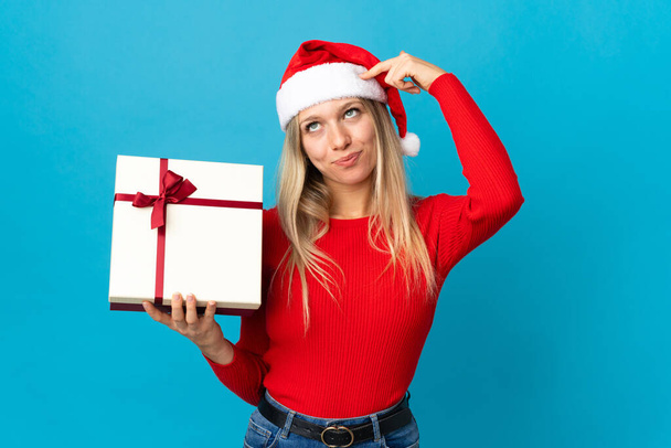 Noel şapkalı bir kadın mavi arka planda izole edilmiş bir hediye tutuyor. Şüpheleri var ve kafası karışmış bir yüz ifadesi var. - Fotoğraf, Görsel