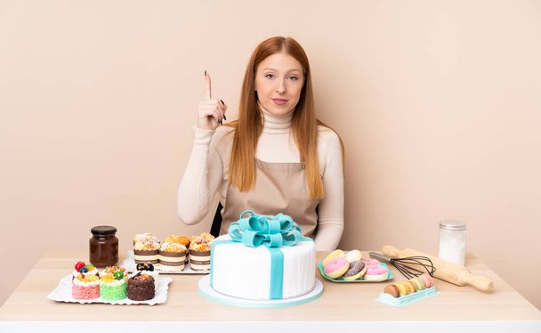 Jeune rousse femme avec un gros gâteau pointant avec l'index une excellente idée - Photo, image