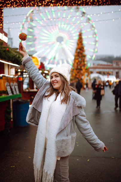 Happy woman on Christmas market with caramelized apple. Outdoor activities on Christmas and New Year. Young woman enjoying winter moments. Lights around. - Фото, зображення