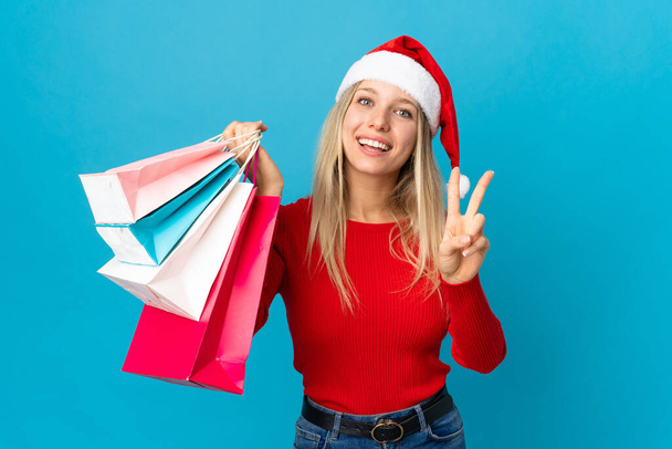 Frau mit Weihnachtsmütze kauft etwas Vereinzeltes auf blauem Hintergrund, lächelt und zeigt Siegeszeichen - Foto, Bild