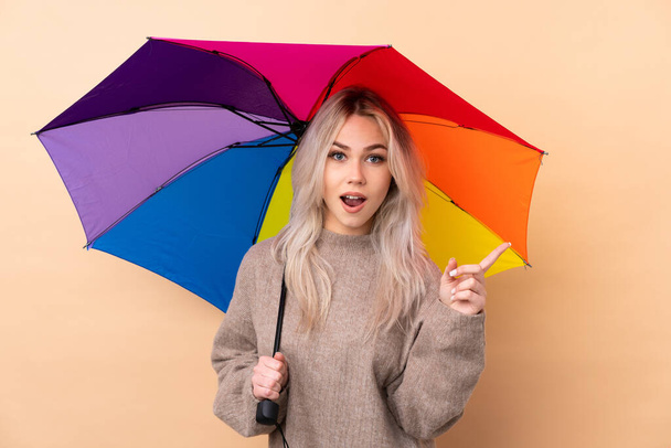 Nastolatka trzymając parasol na odosobnionym tle zaskoczony i wskazując palcem w bok - Zdjęcie, obraz
