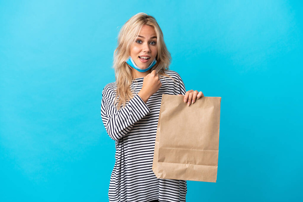 Young Russian woman holding a grocery shopping bag isolated on blue background celebrating a victory - Φωτογραφία, εικόνα