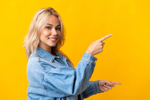 側に指を指して製品を提示黄色の背景に隔離された若いロシアの女性 - 写真・画像