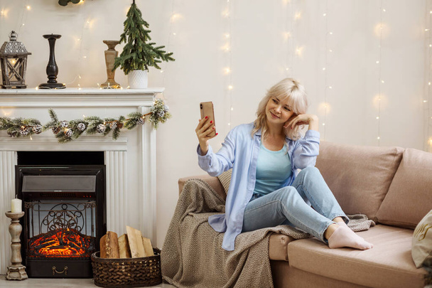 Attractive woman is taking selfie while sitting on the sofa next to the fireplace. Celebrating Christmas at home, cosy interior.  - Foto, Imagen
