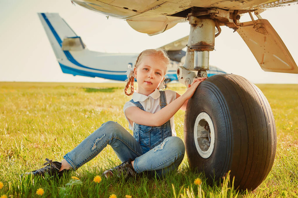 Cute little girl sitting near the landing gear wheels of an airplane and dreaming about traveling and flying in an airplane. - Foto, Imagem