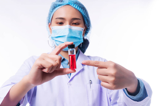 Hand of young woman doctor wear uniform white clothes and medical mask protection virus disease on nose while holding use finger pointing to test tube with blood sample over isolated white background. Select focus - Photo, Image