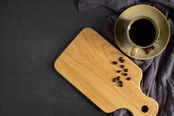 Top view above of Black hot fresh coffee and wooden chopping board with handle and hole for hanging and coffee beans roasted, apron on dark black table background. with copy space. - Foto, Imagem