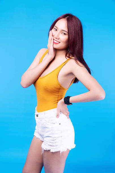 Cheerful young woman brown long hair wear short jeans holding hand on her face while standing over isolated blue background. Positive girl smiling posture while looking at camera with copy space. - Photo, Image