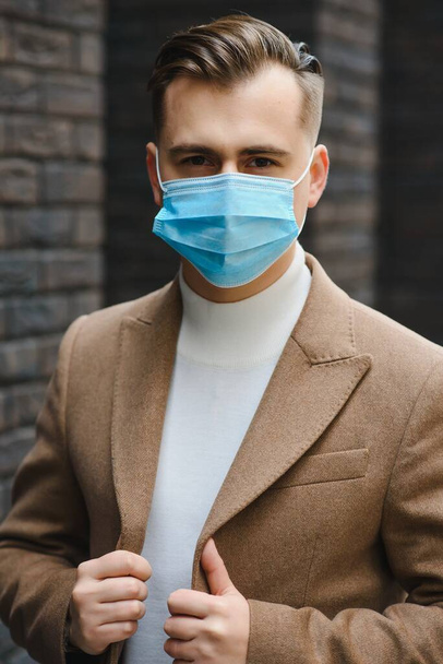Businessman and coronavirus epidemic. Close up portrait of young business man in a disposable facial mask. A man defends himself against covid 19 on the big city street. Modern buildings at background - Фото, изображение