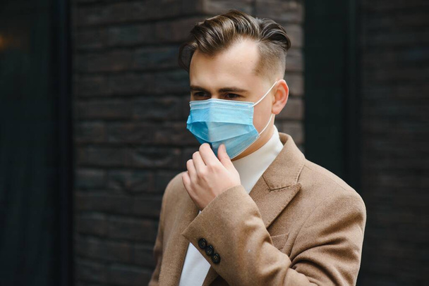 Businessman and coronavirus epidemic. Close up portrait of young business man in a disposable facial mask. A man defends himself against covid 19 on the big city street. Modern buildings at background - Zdjęcie, obraz