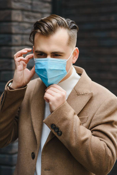 Businessman and coronavirus epidemic. Close up portrait of young business man in a disposable facial mask. A man defends himself against covid 19 on the big city street. Modern buildings at background - Photo, image