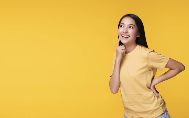 Wesoła młoda kobieta używa palca wskazującego jej twarz pustą kopią przestrzeni stojąc nad odizolowanym na żółtym tle - Zdjęcie, obraz