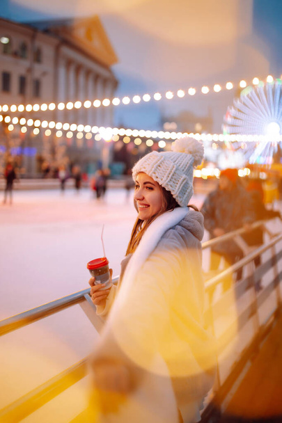 Happy young woman on Christmas market with coffe near skating rink. Outdoor activities on  Christmas and New Year. Lights around.Winter holiday. - Фото, зображення